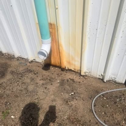 White metal wall with rust stain, turquoise downspout, and muddy ground beside a hose.