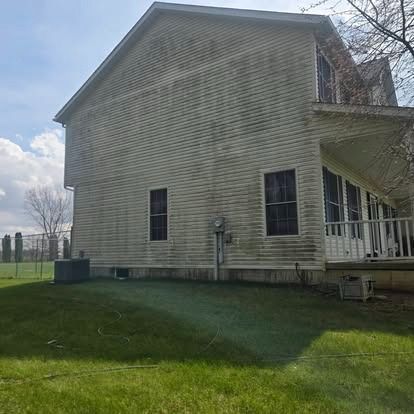 Side view of a beige house with a porch and green lawn under a partly cloudy sky