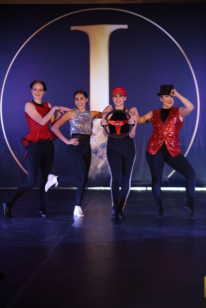 Four performers in glittering costumes dance on stage before a large illuminated letter I.