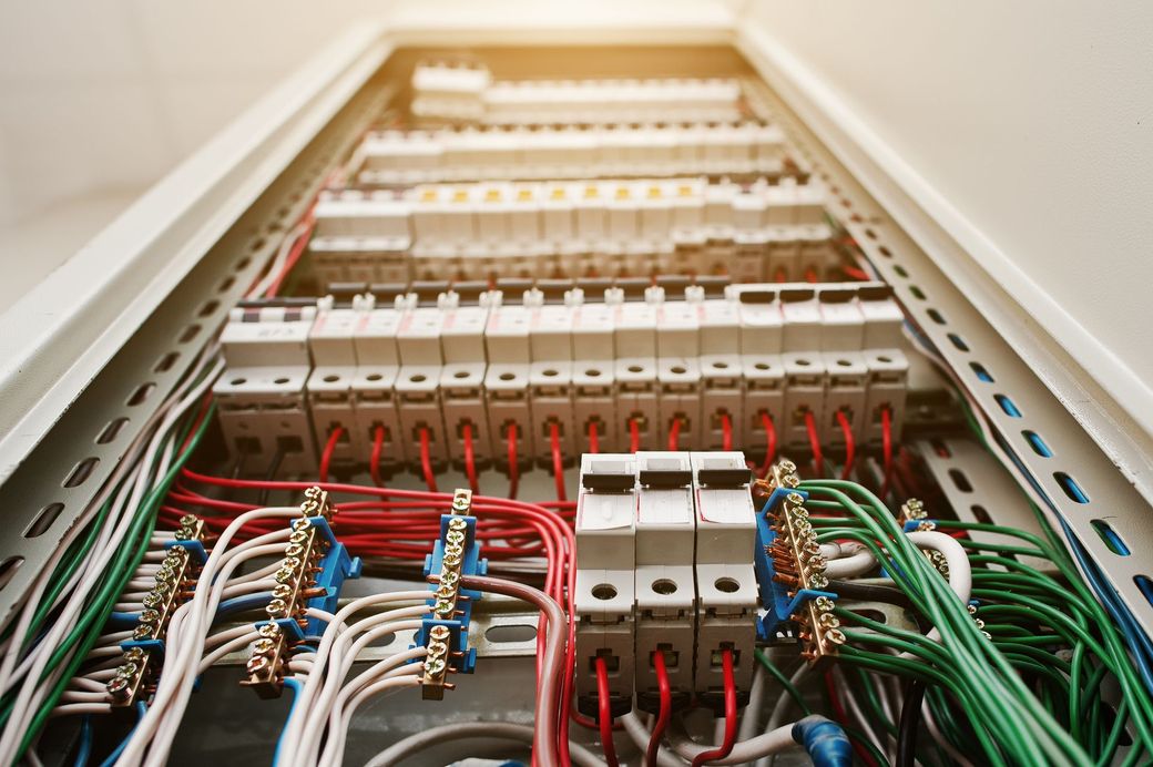 An open electrical panel with rows of white circuit breakers, red wiring, and bundles of white and green grounding wires.