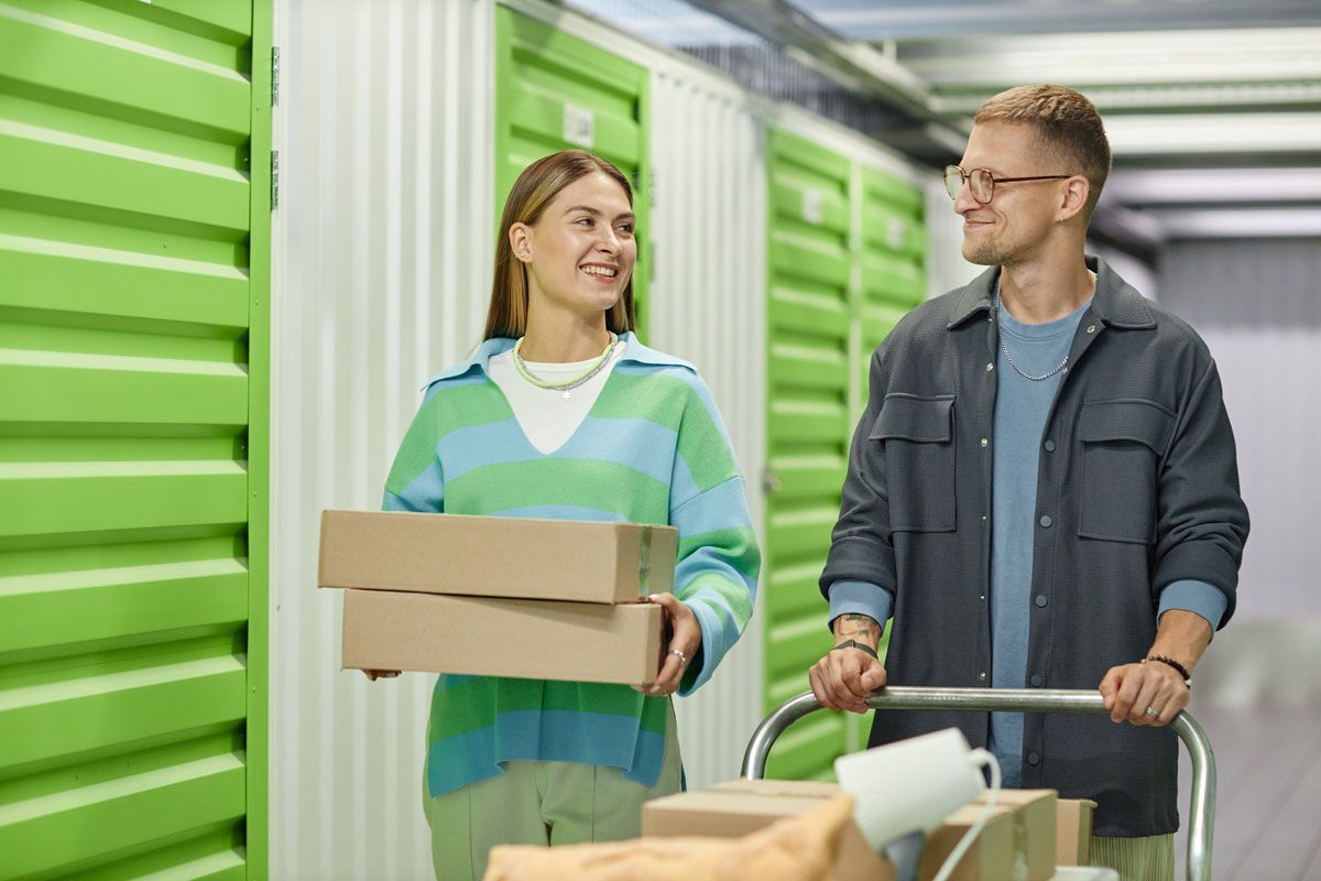 Two people with luggage carts in a green storage facility, one carrying a box and smiling.