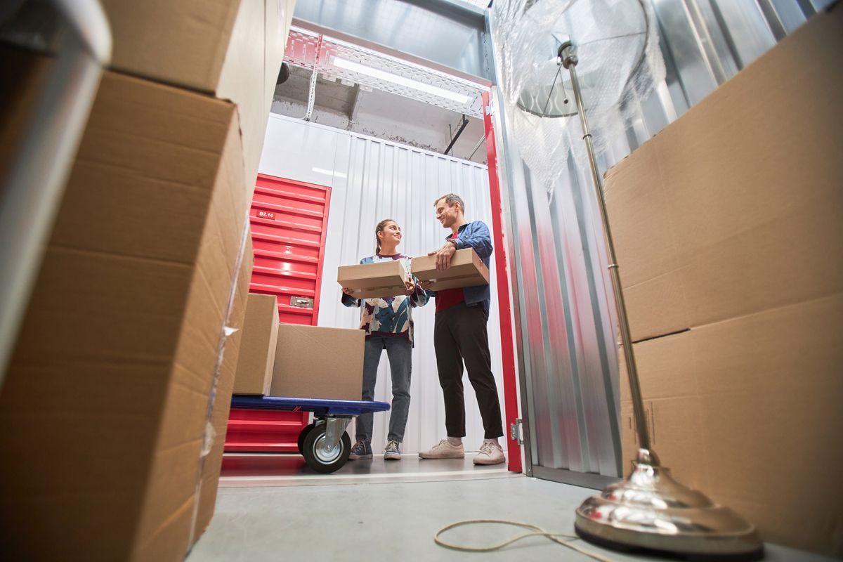 Two workers carrying a box between cardboard storage stacks in a warehouse aisle