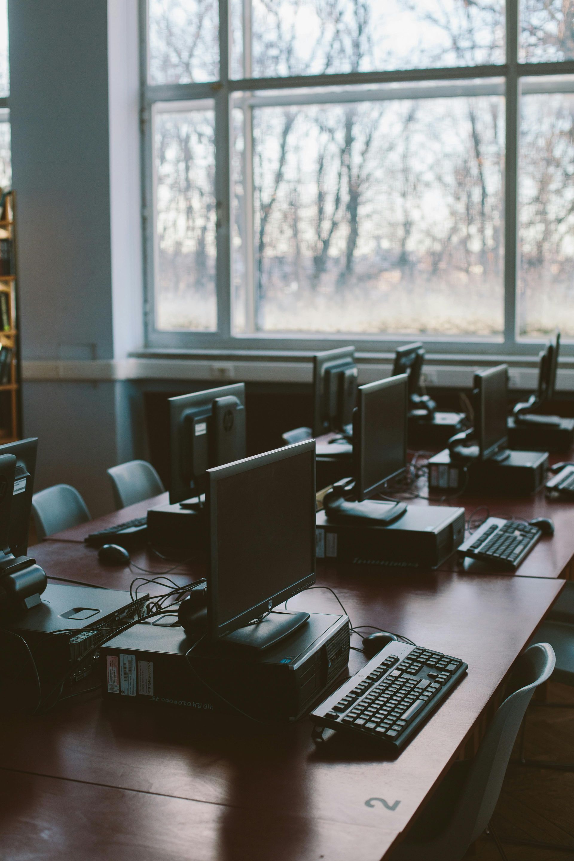 Salle informatique avec des rangées d'ordinateurs sur une longue table en bois près d'une grande fenêtre