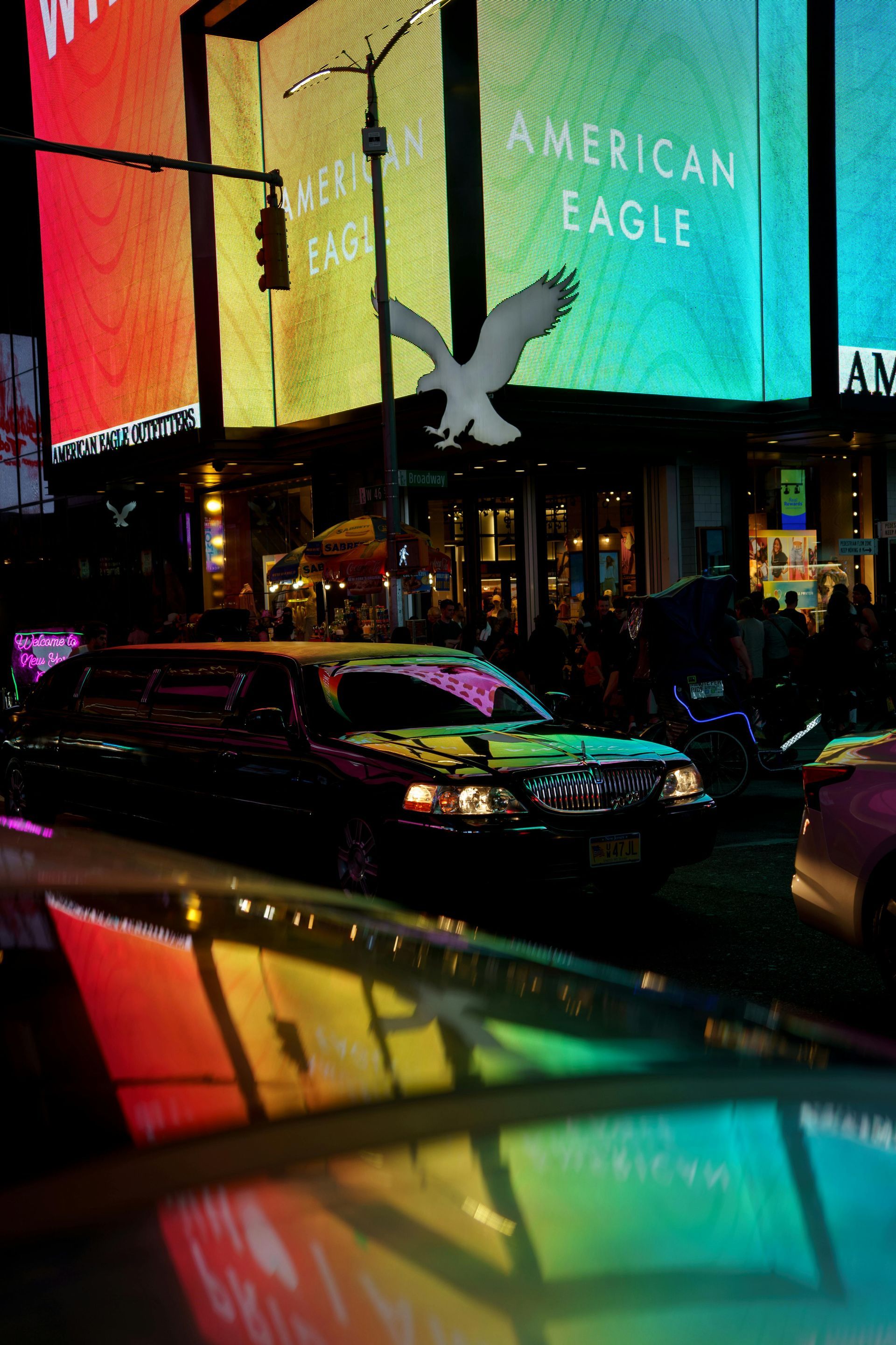 A limousine in Times Square at night, with neon-lit American Eagle sign. Reflections on car hoods.
