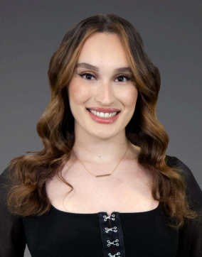 Smiling person with wavy brown hair wearing a black top against a gray background