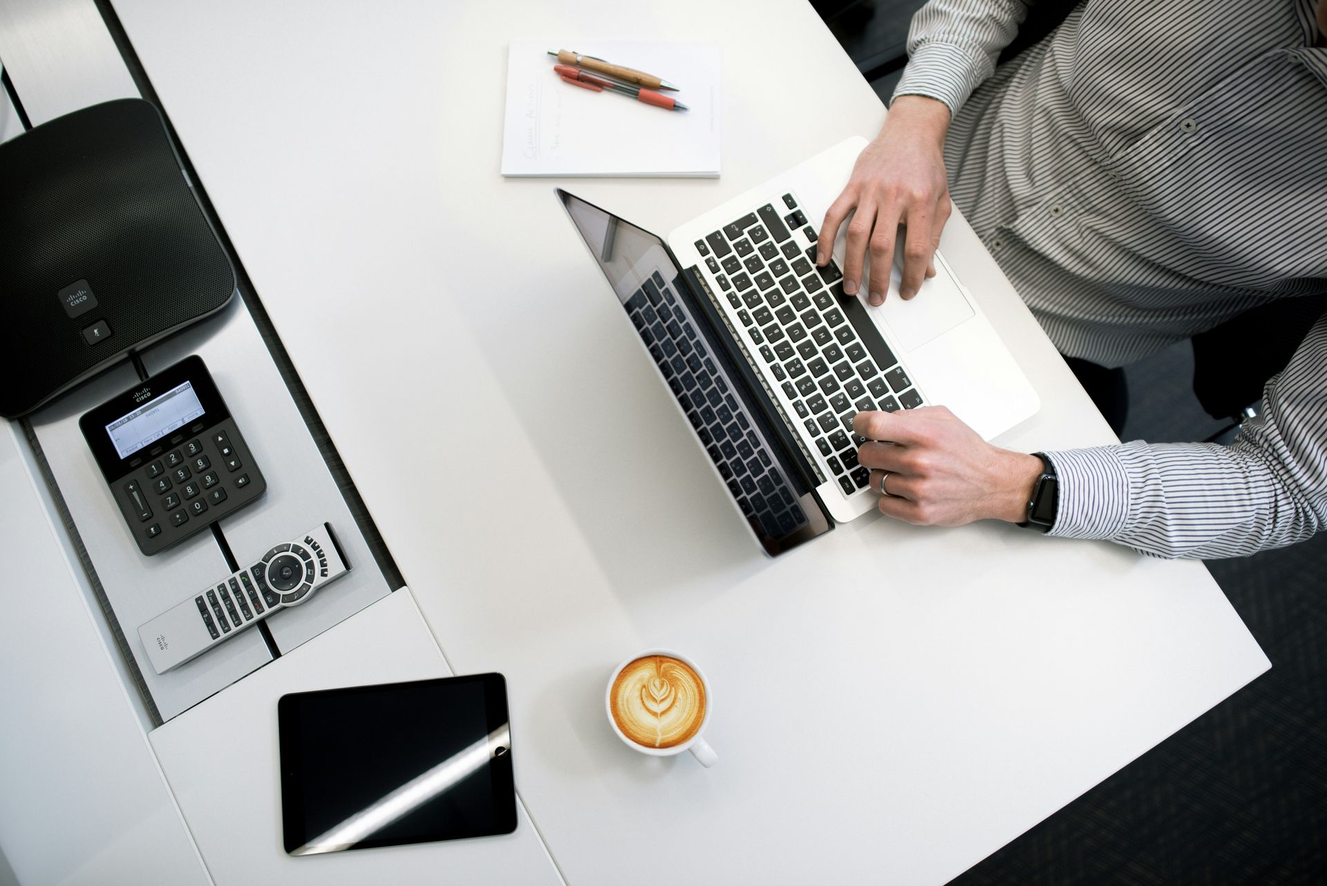 Person typing on a laptop at a white table with a coffee cup, tablet, and office equipment.