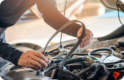 A mechanic holds a serpentine belt while working on an open car engine in a well-lit workshop.