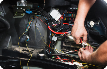 A close-up of a person's hands using pliers to strip wires near a vehicle's open fuse box and electrical wiring.