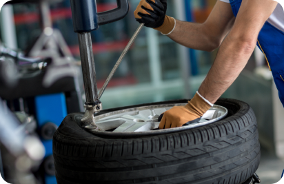 A mechanic in blue coveralls uses a tire changer machine to mount a tire onto a car wheel rim in a repair shop.