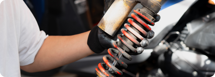 A mechanic in a white shirt and black glove holds a metal motorcycle shock absorber with a rusted red coil spring.
