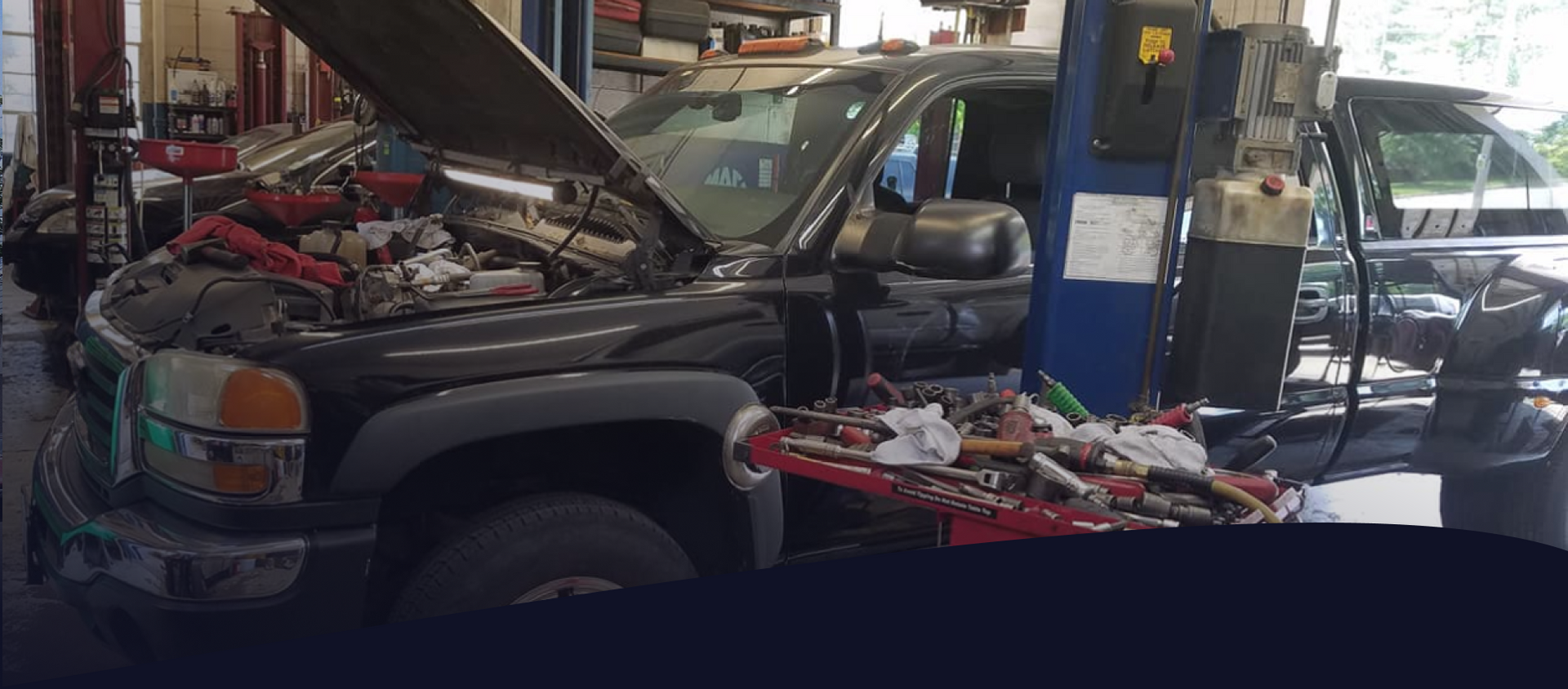 A black pickup truck with its hood open, elevated on a car lift inside an auto repair shop with tools nearby.