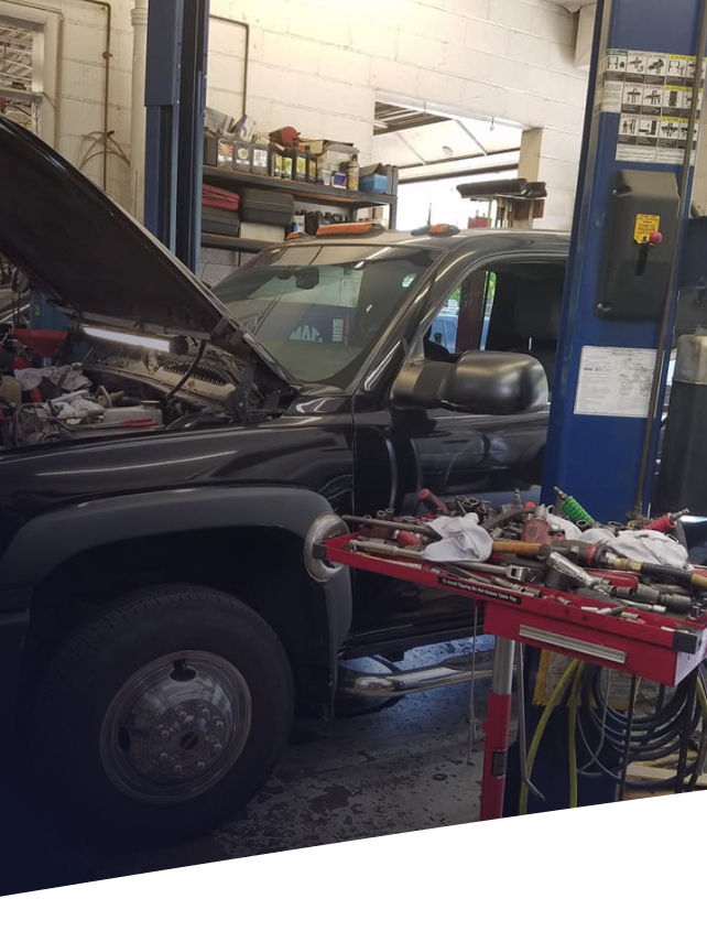 A black pickup truck with its hood open, elevated on a car lift inside an auto repair shop with tools nearby.