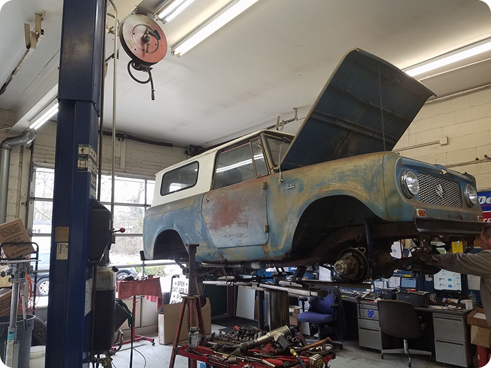 A weathered blue and white vintage SUV sits elevated on a lift inside a mechanic's garage with its hood open.