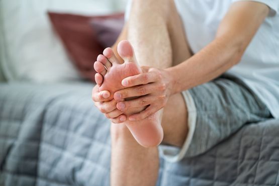 A person sitting on a bed, holding and rubbing their bare foot with both hands, showing signs of foot pain.