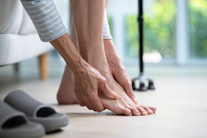 A person sitting down, holding and rubbing their sore foot. A cane and pair of slippers are nearby.