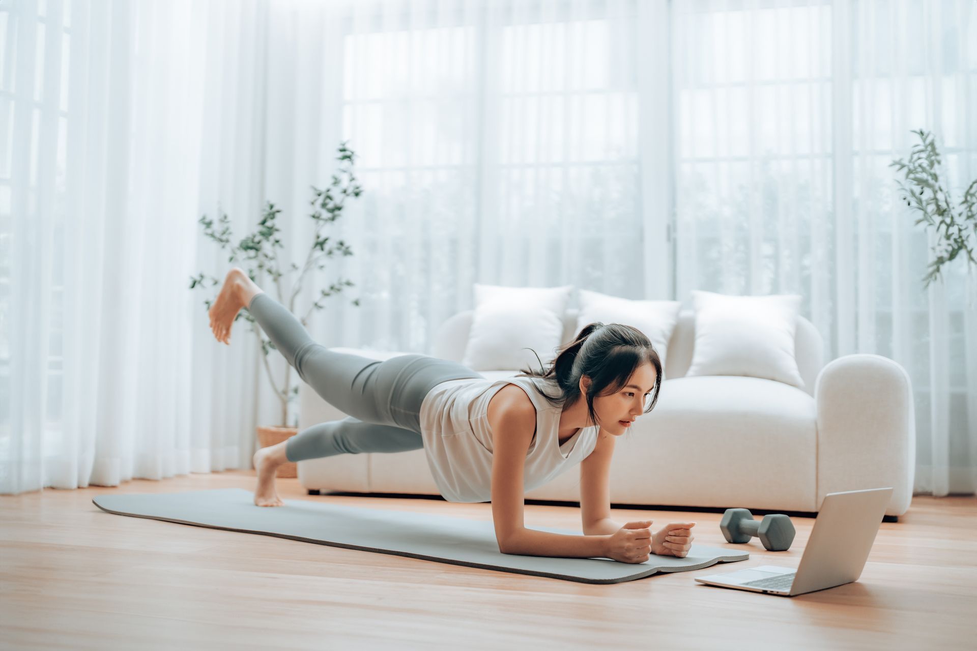 Person doing a forearm plank workout on a yoga mat in a bright living room, facing a laptop.
