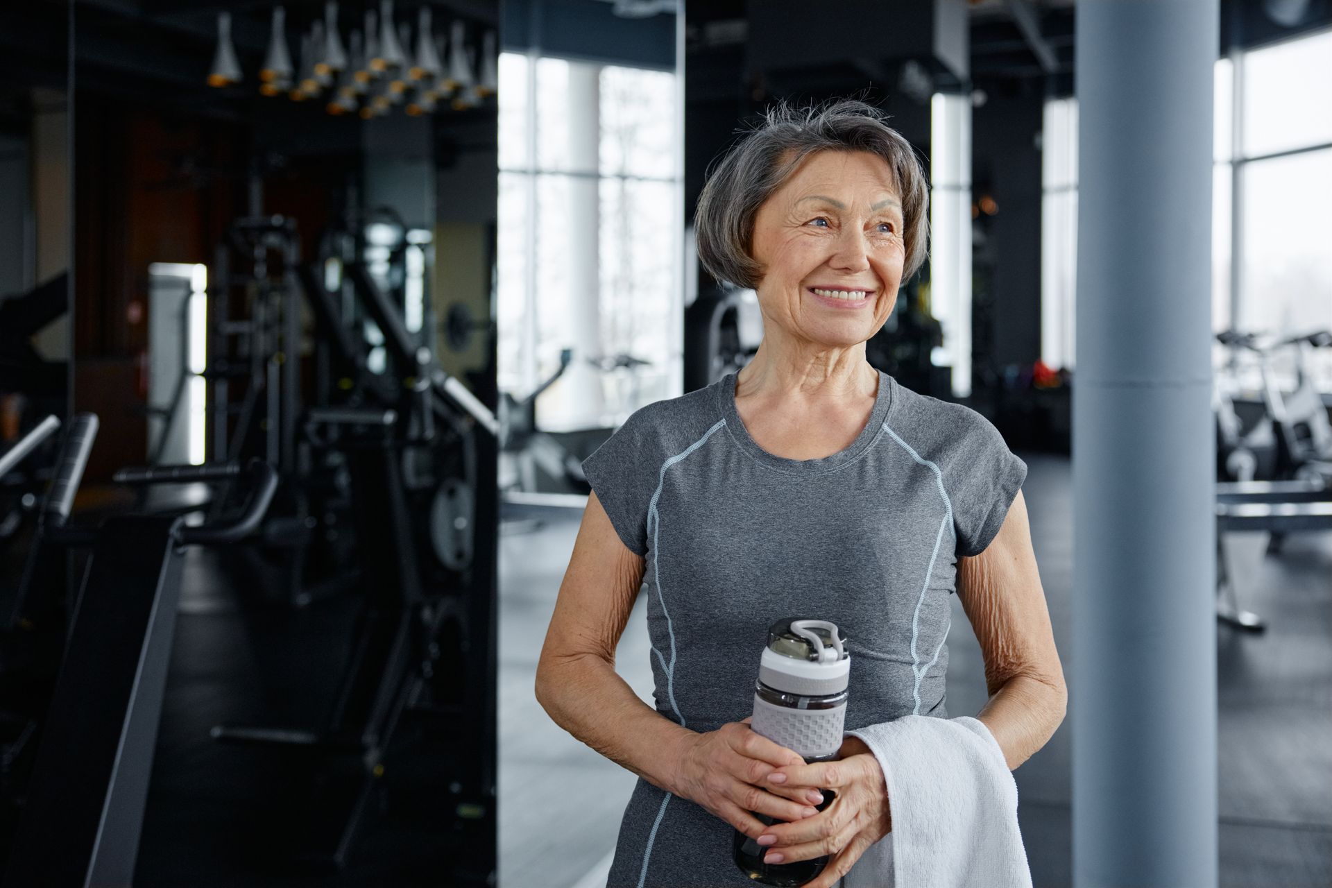 Smiling woman in a gym holding a water bottle and towel with headphones on