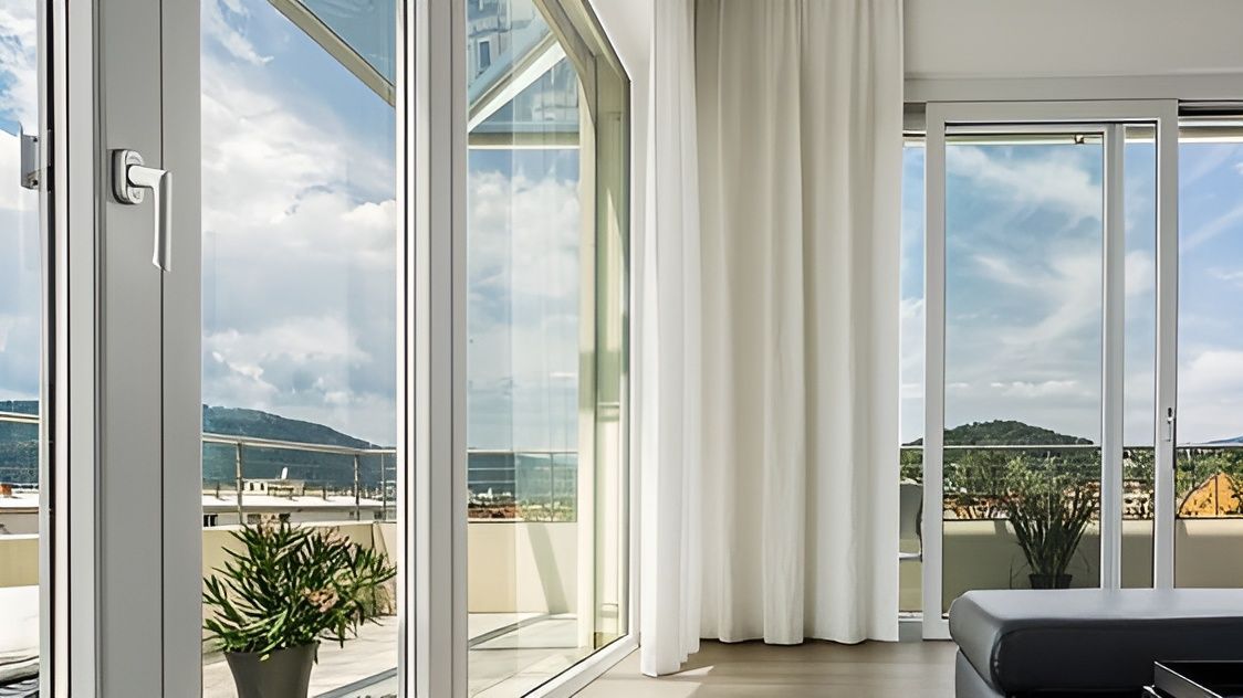 Luminoso soggiorno con porte finestre che si aprono sul balcone, tende bianche e vista sulle colline e sul mare.