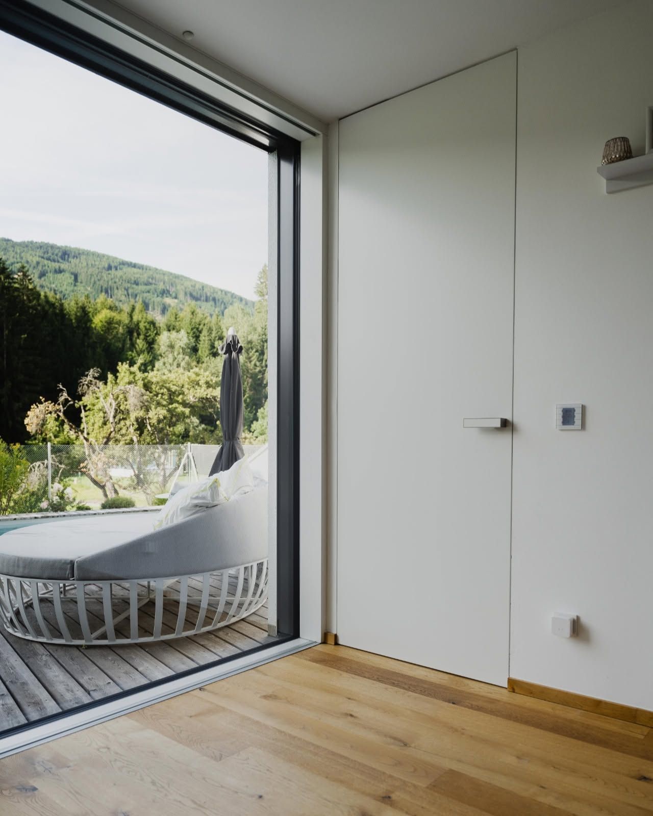 Camera moderna con porta scorrevole che si apre su un balcone con vista sulle montagne, pavimento in legno chiaro, pareti bianche e sedia grigia.