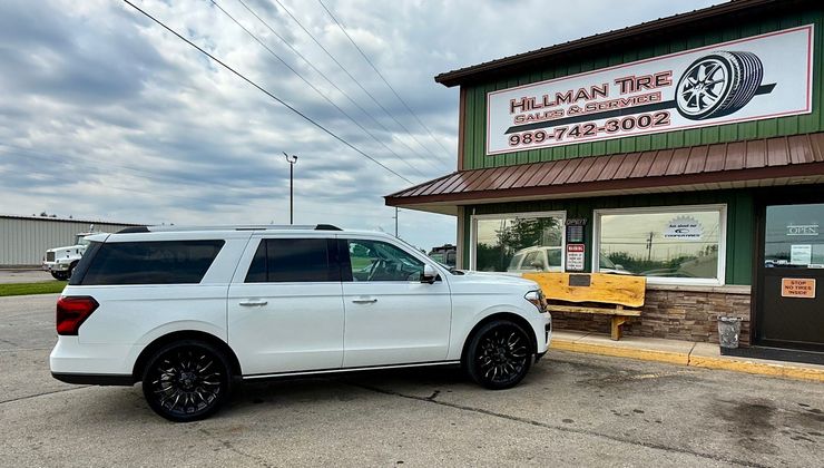 A white SUV parked in front of a building with a "Hillman Tire & Automotive" sign on a cloudy day. | Hillman Tire Sales and Service LLC