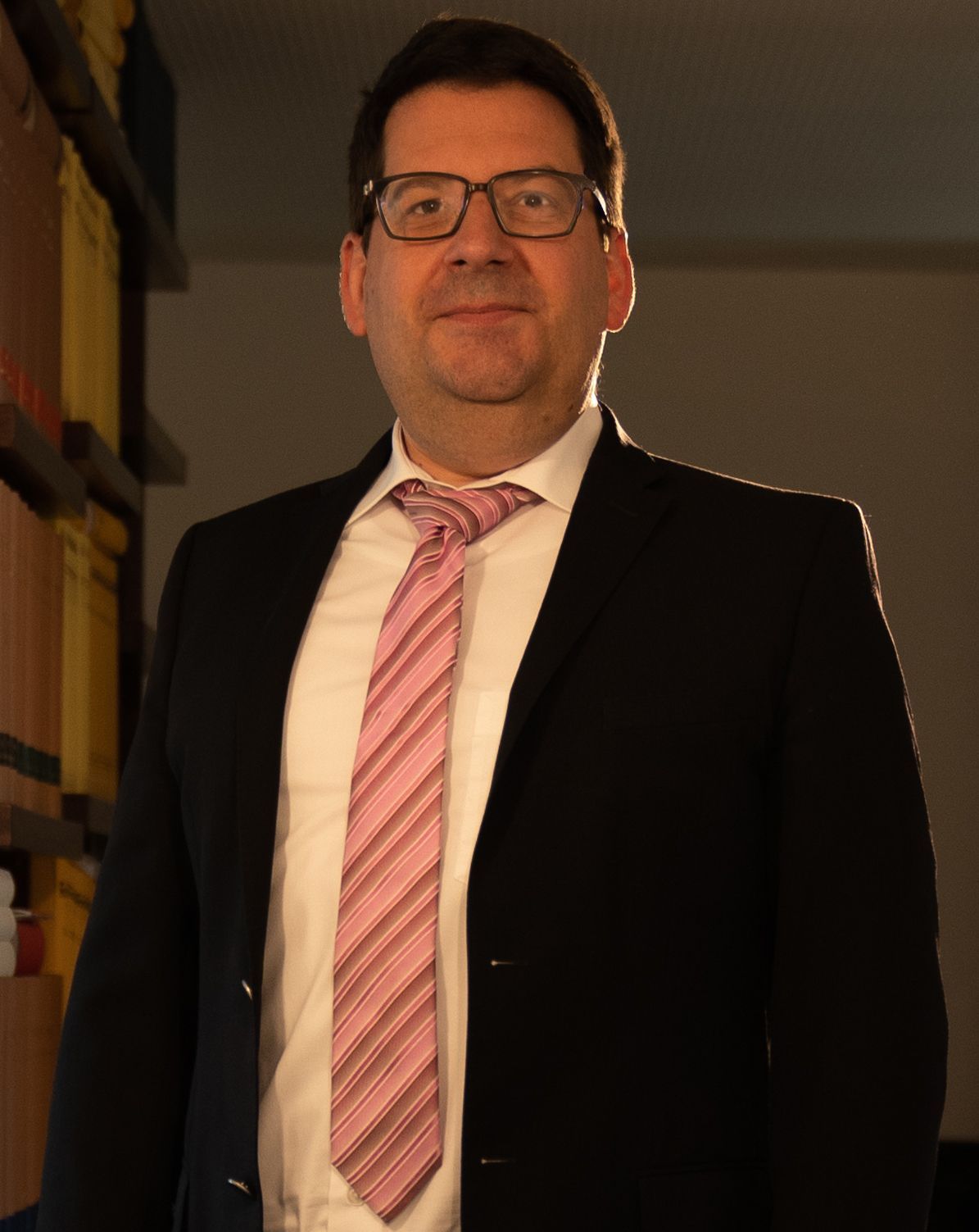 A man in a suit and tie is standing in front of a bookshelf.