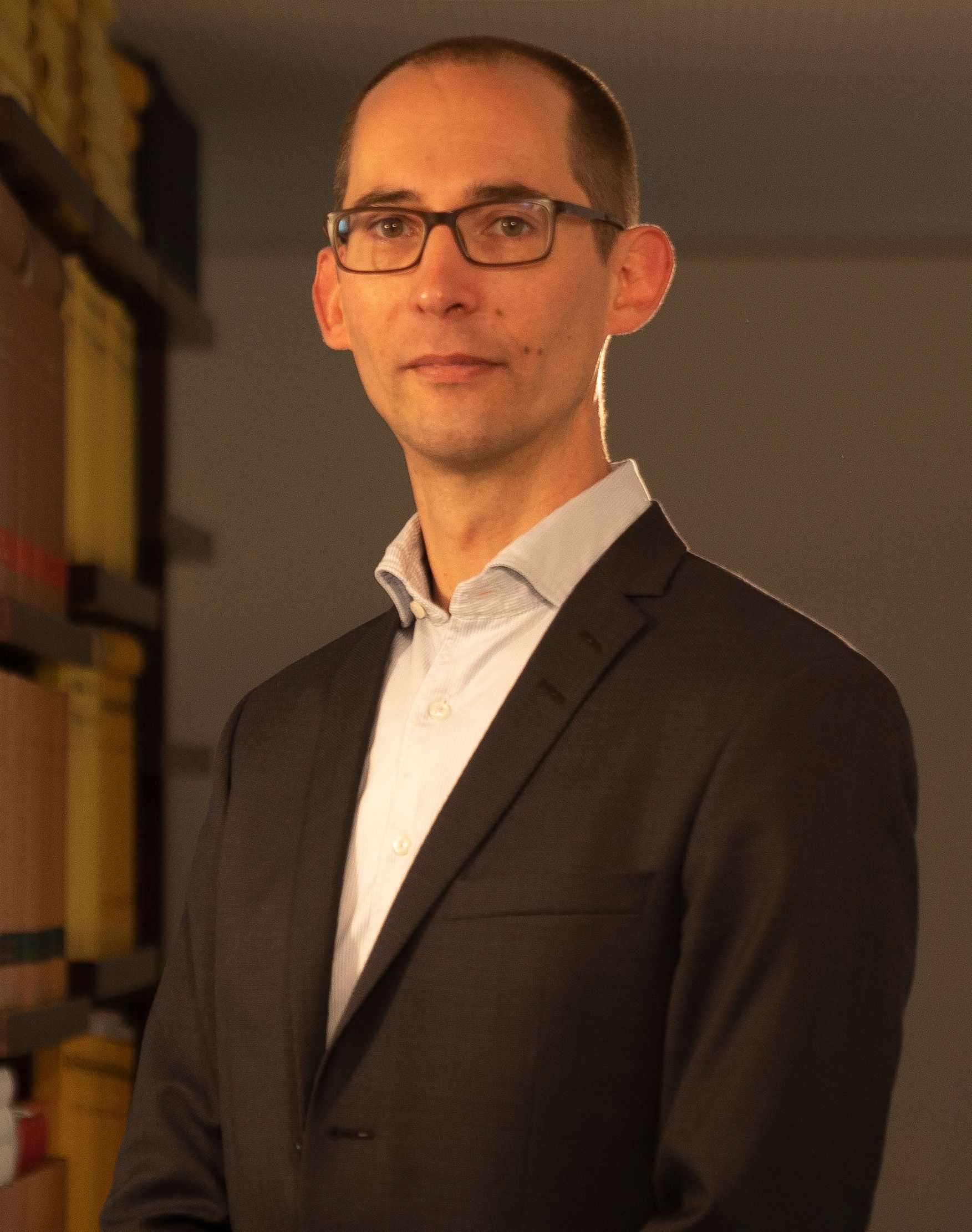 A man in a suit and glasses is standing in front of a bookshelf.