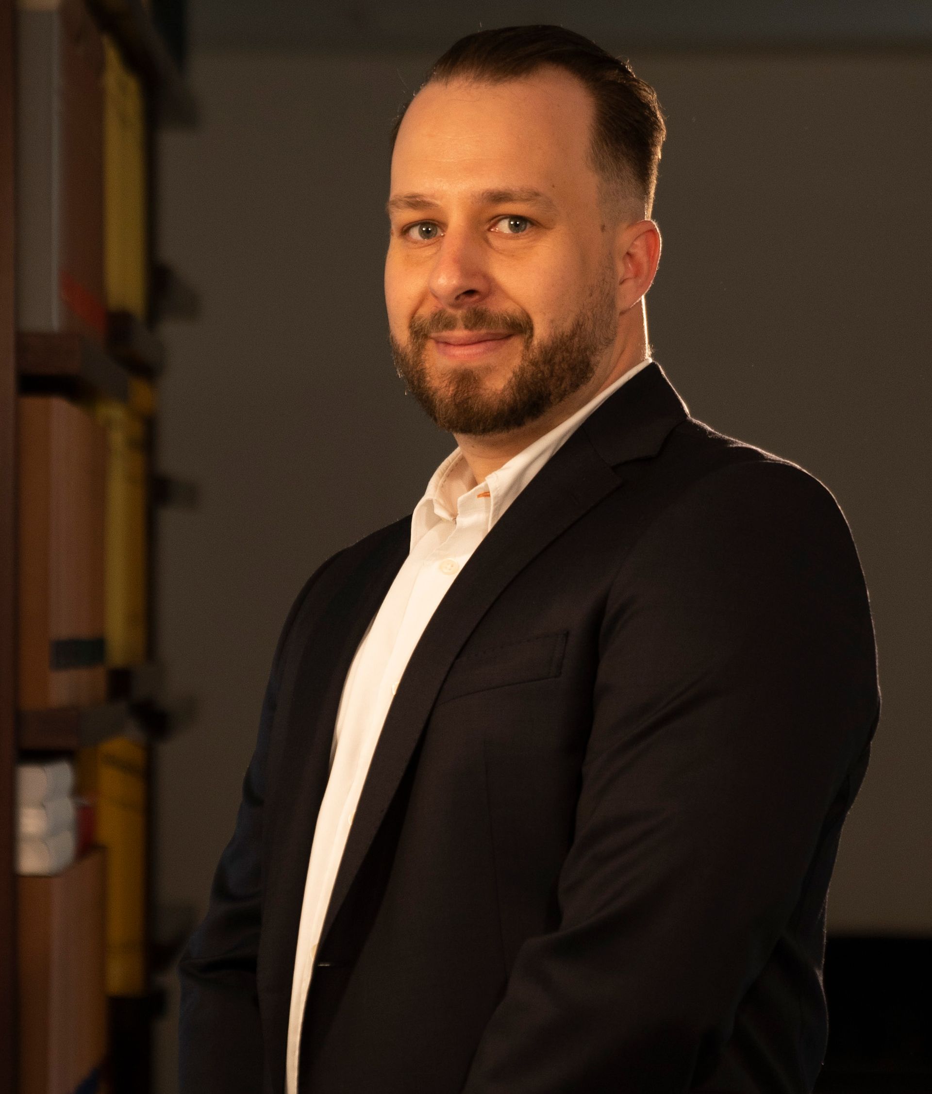 A man in a suit and white shirt is standing in front of a bookshelf.