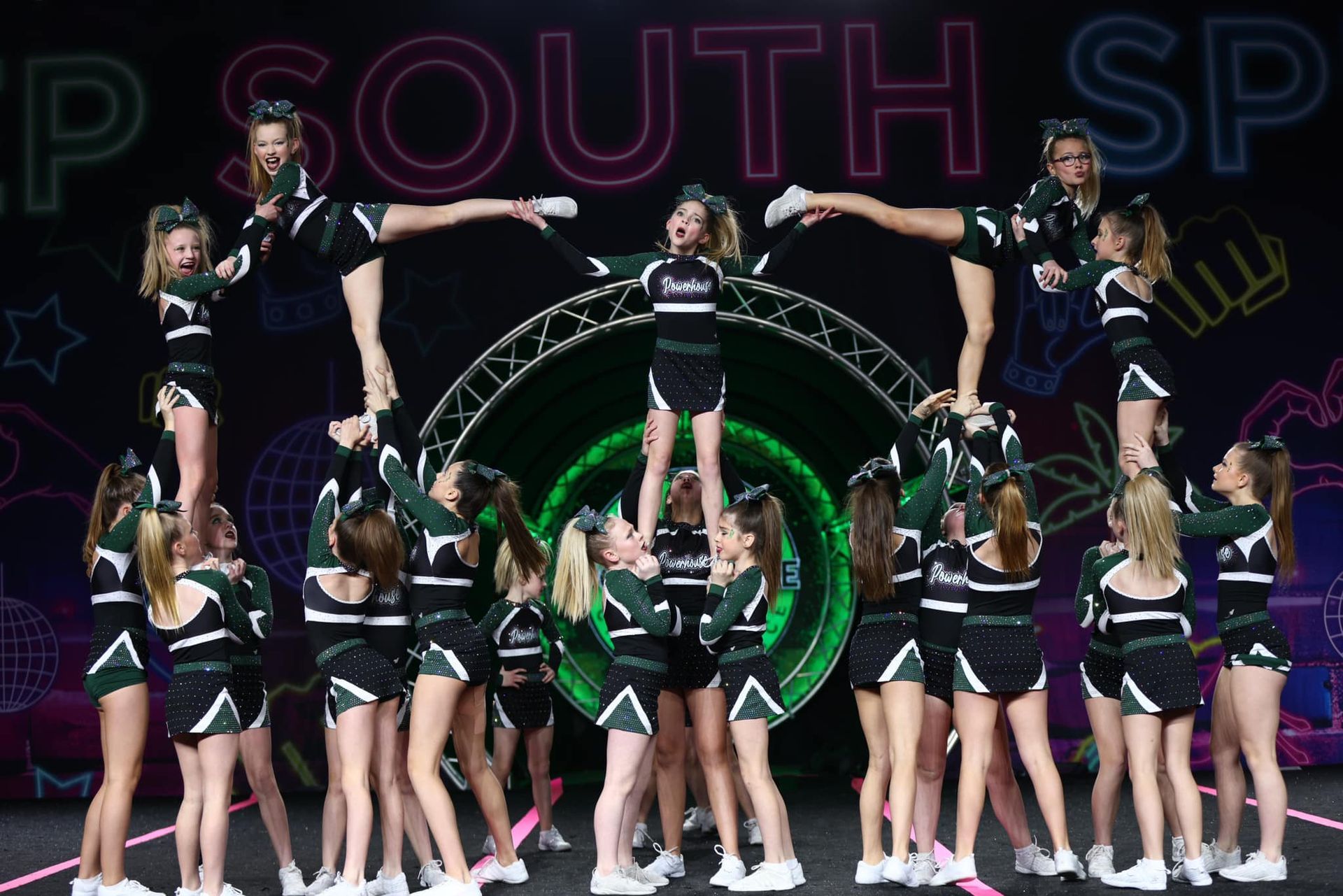 Cheerleaders performing a pyramid stunt on a competition mat with a neon green arch backdrop