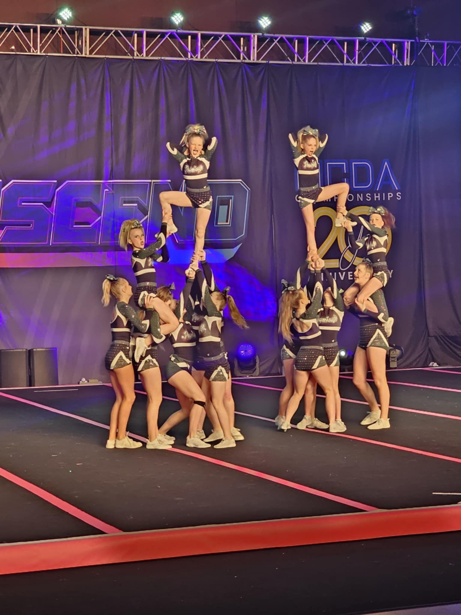 Cheerleading team building a multi-level stunt on a lit competition stage with purple curtains