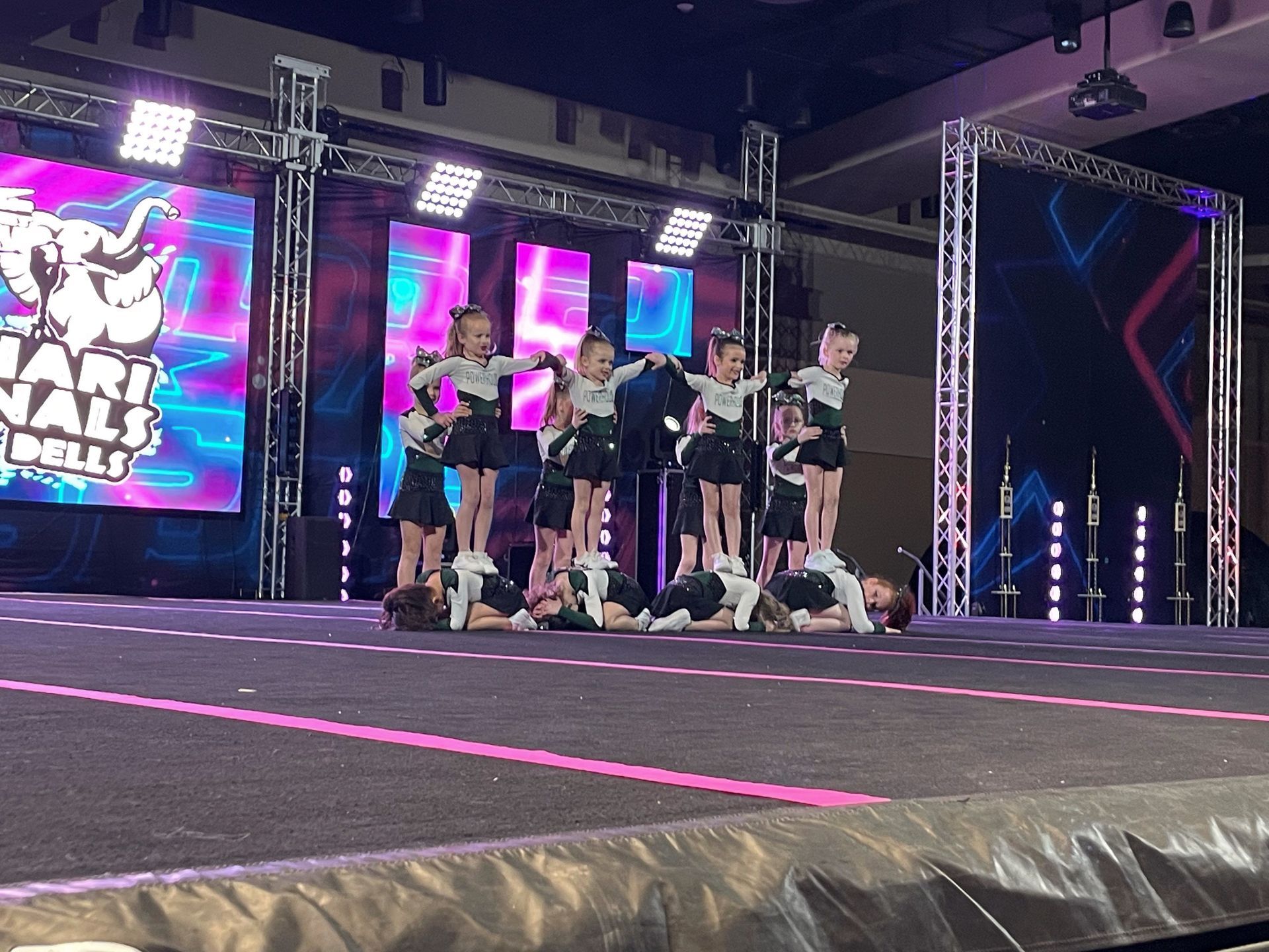 Cheer team posing on a lit stage with pink accents and a large event screen in the background