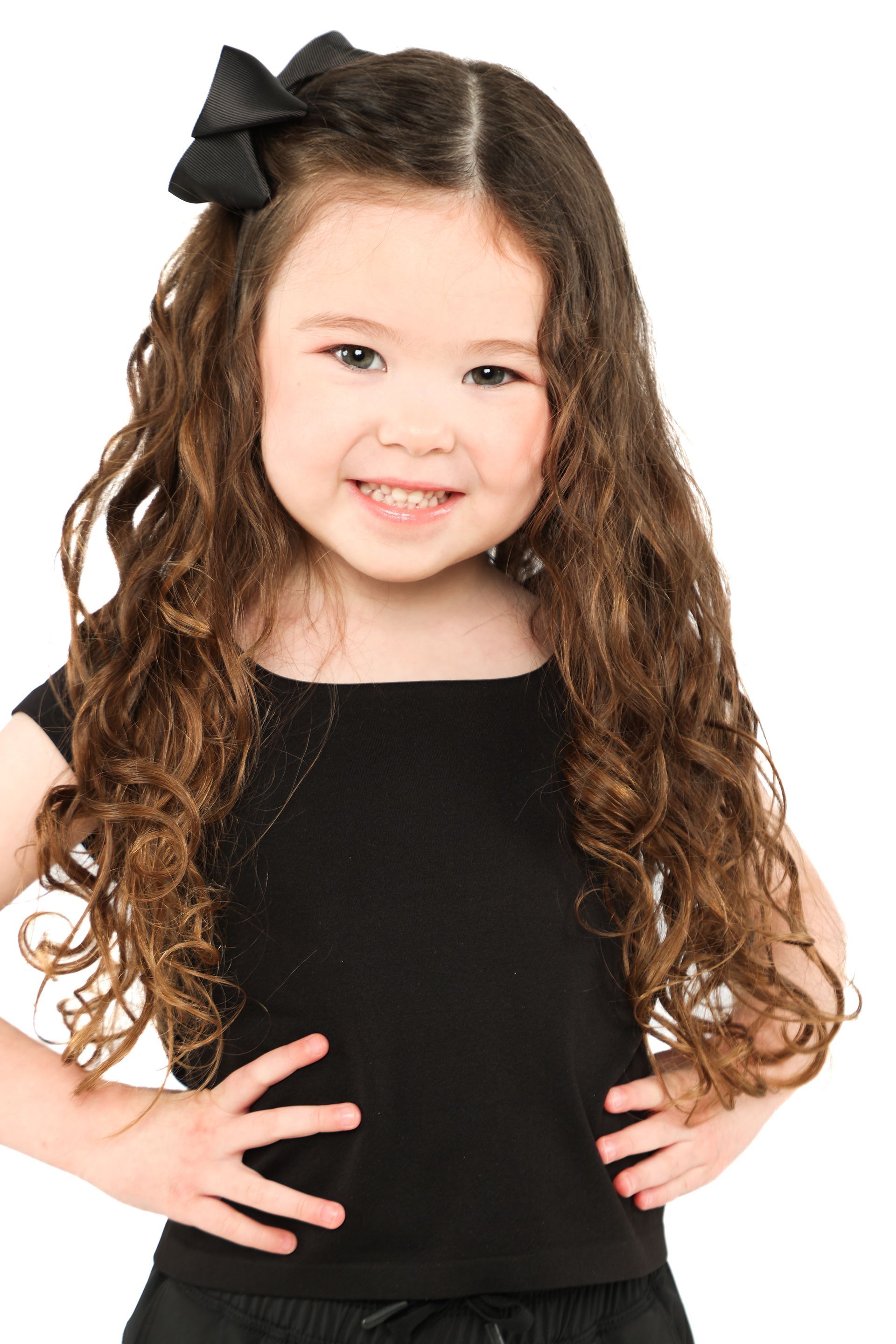 Smiling child in a black dress with a black bow in her curly hair, hands on hips, against a white background