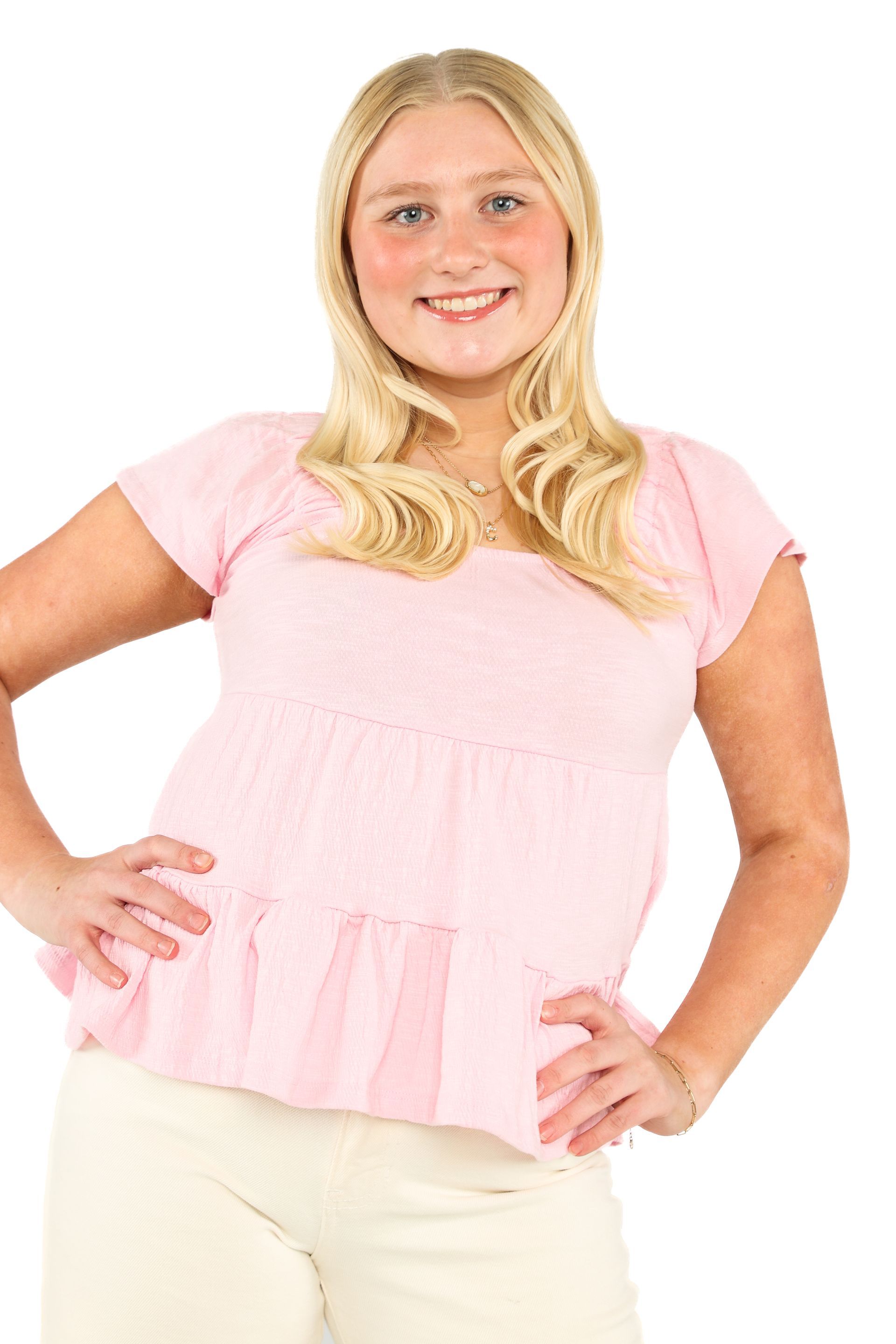Blonde person in a pink top and white pants standing with hands on hips against a white background