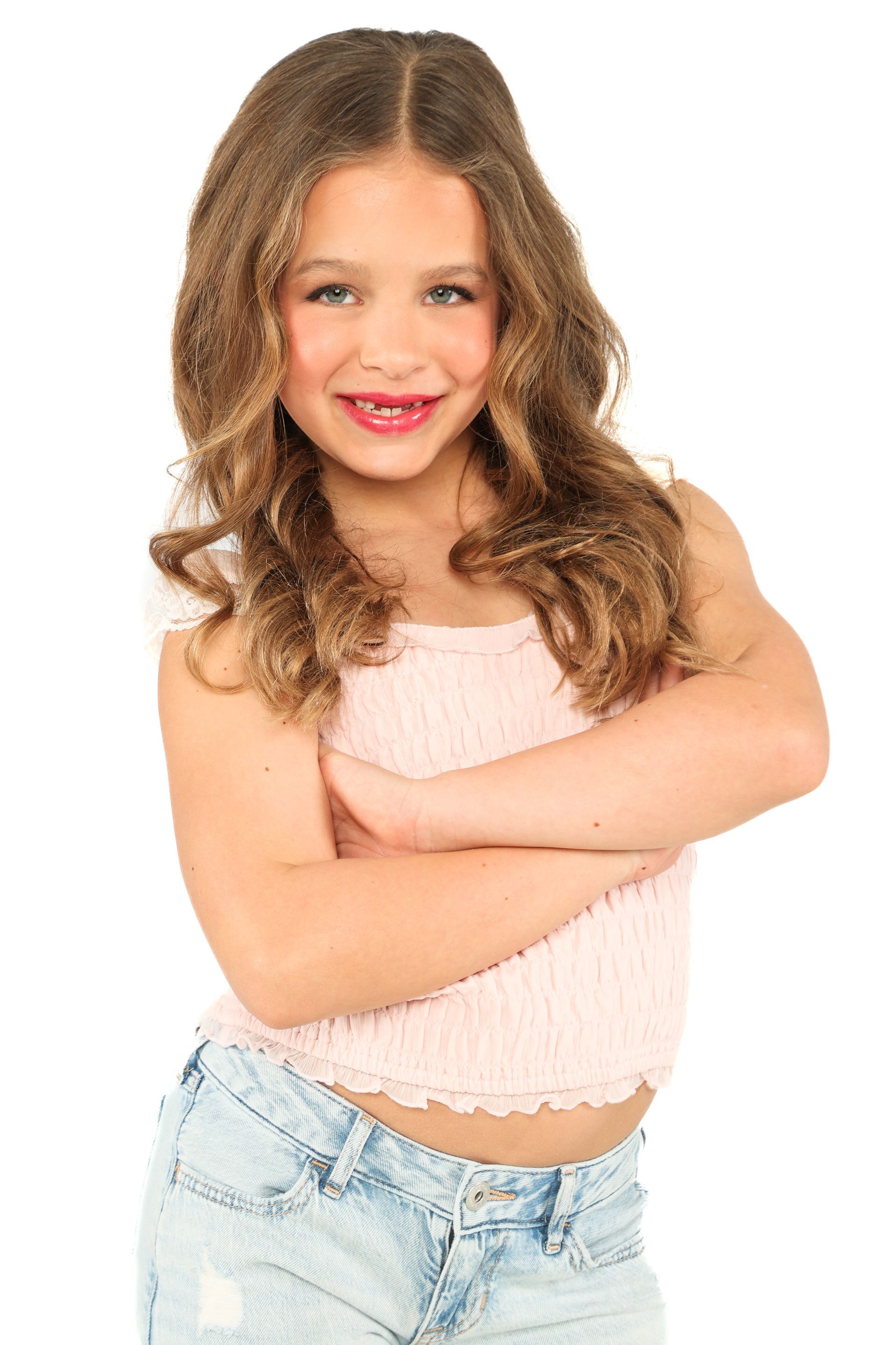 Smiling child with long curly hair, wearing a pink top and light jeans, standing with arms crossed on a white background