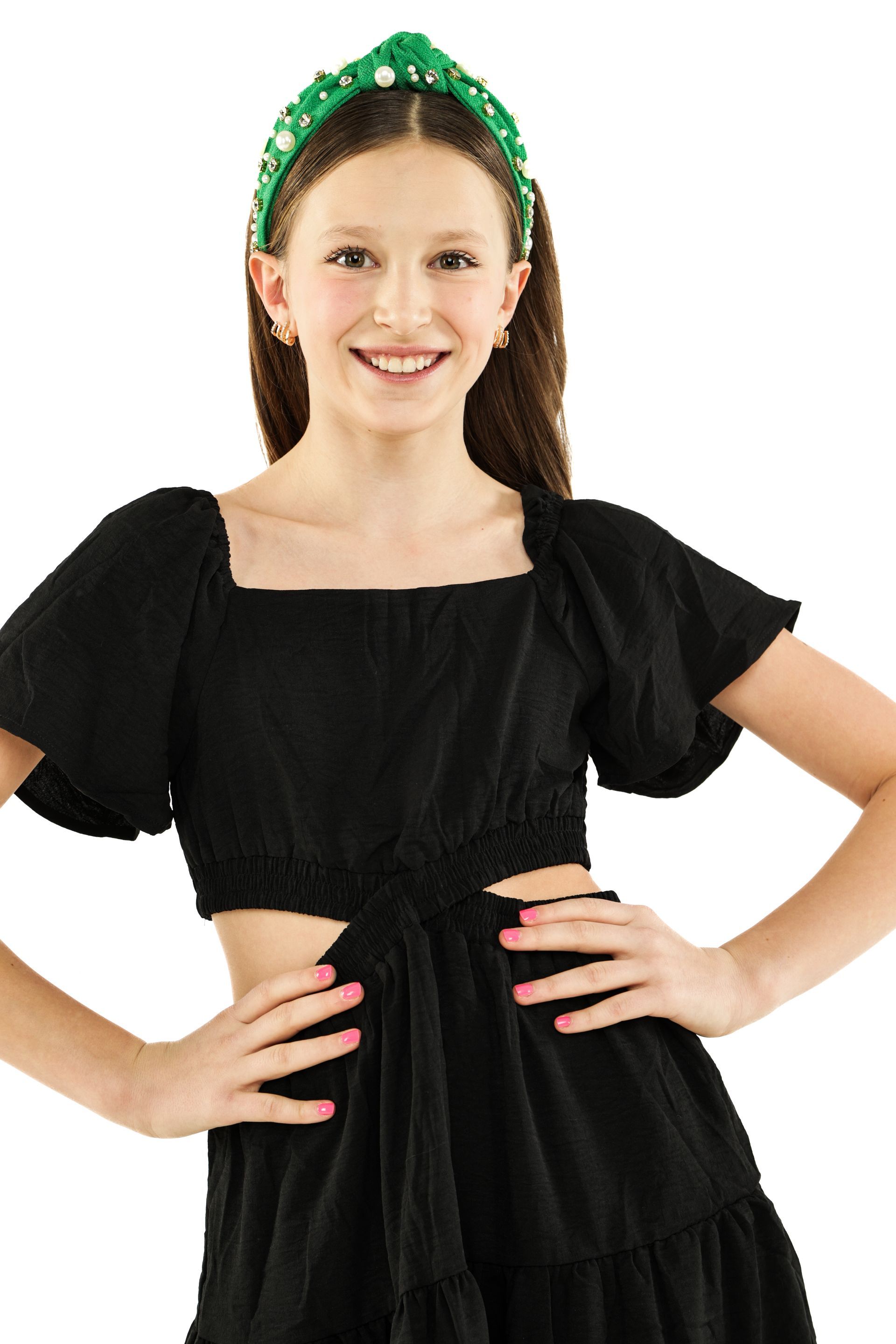 Smiling woman in a black dress and green headband, standing with hands on hips against a white background