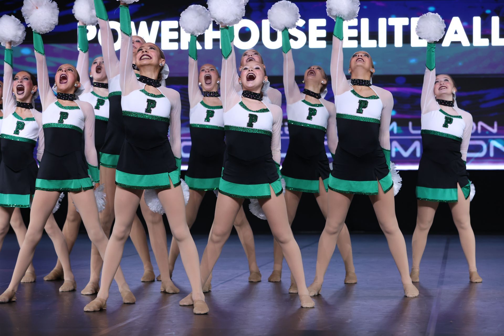 Cheerleaders in black and green uniforms perform a synchronized routine on a stage with arms raised