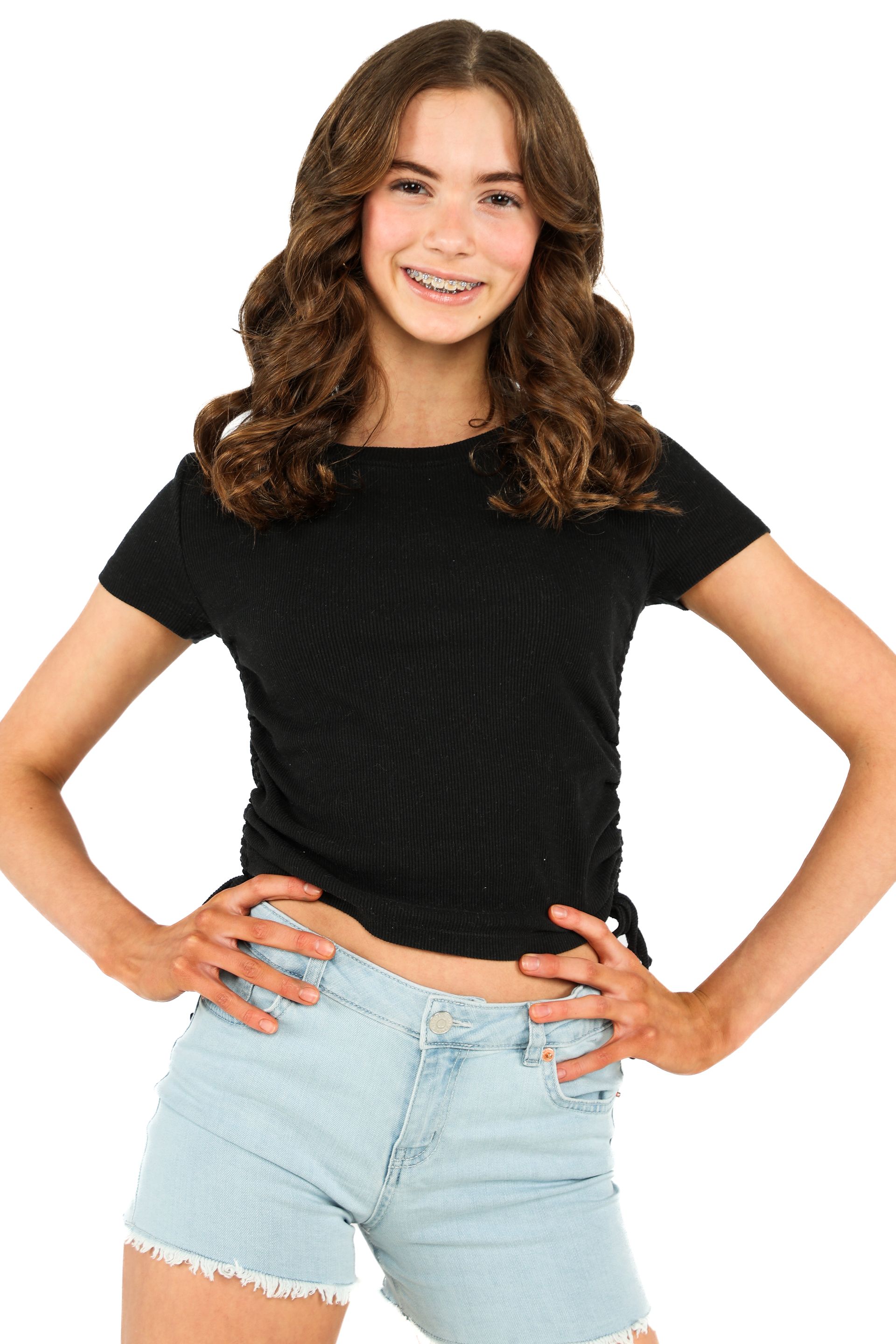 Smiling person in black top and light denim shorts posing with hands on hips against a white background