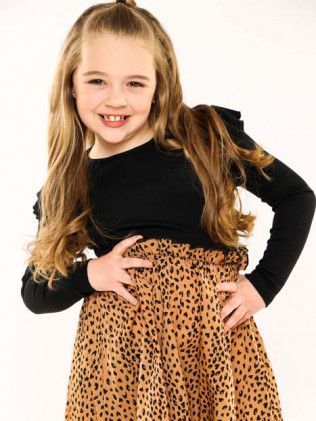 Smiling child in black top and leopard-print skirt, posing with hands on hips against a white background