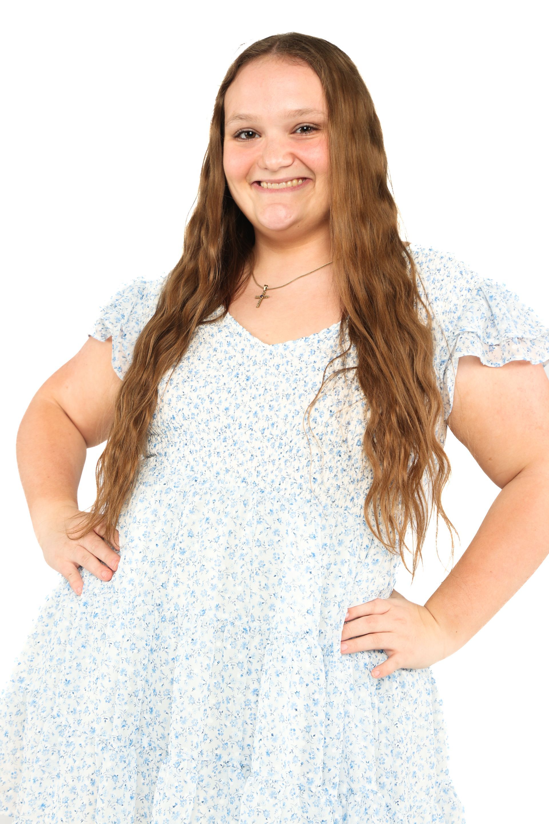 Smiling person in a light blue floral dress, standing with hands on hips against a white background