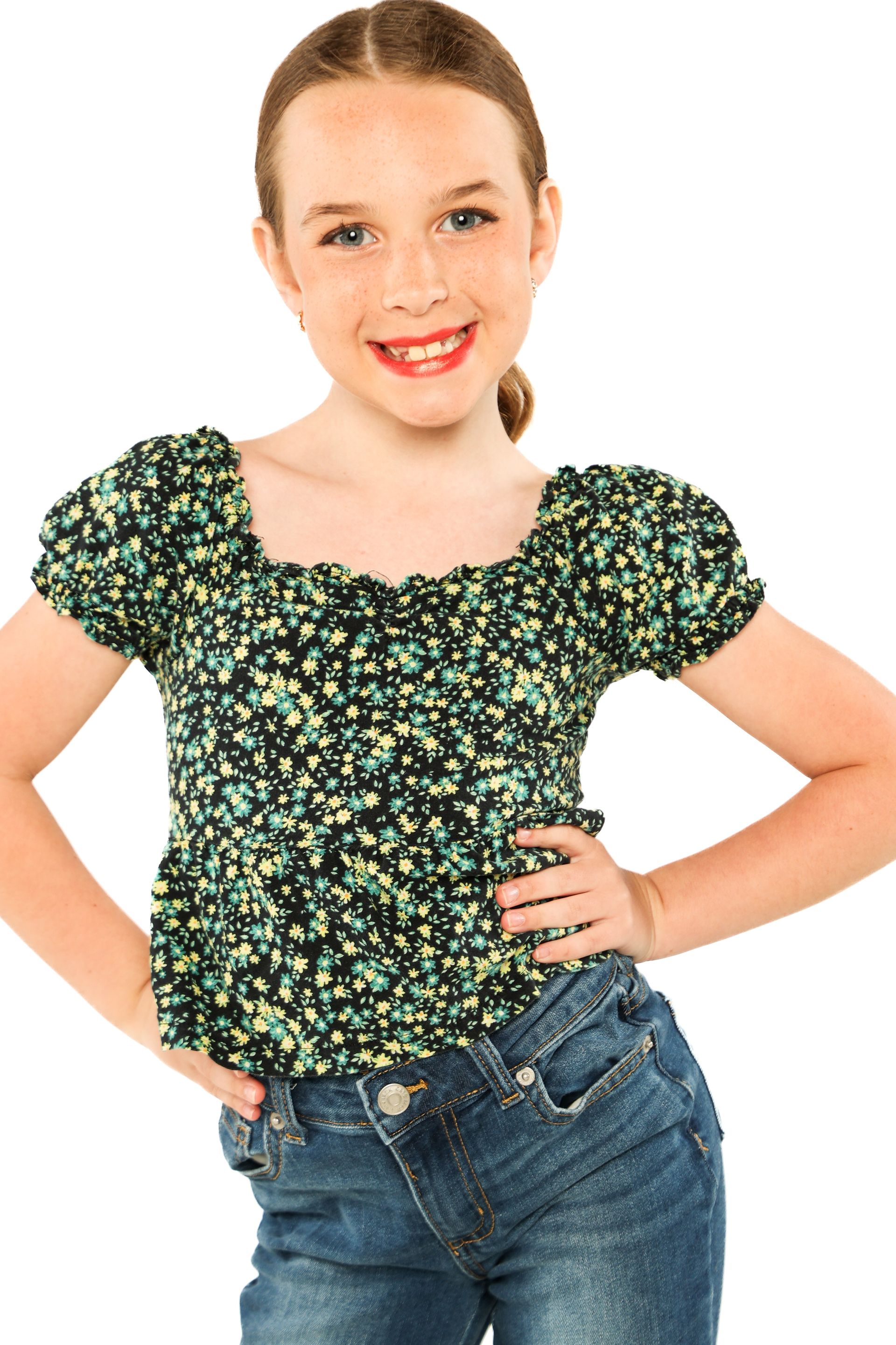 Smiling child in a green floral top and blue jeans, standing with hands on hips against a white background