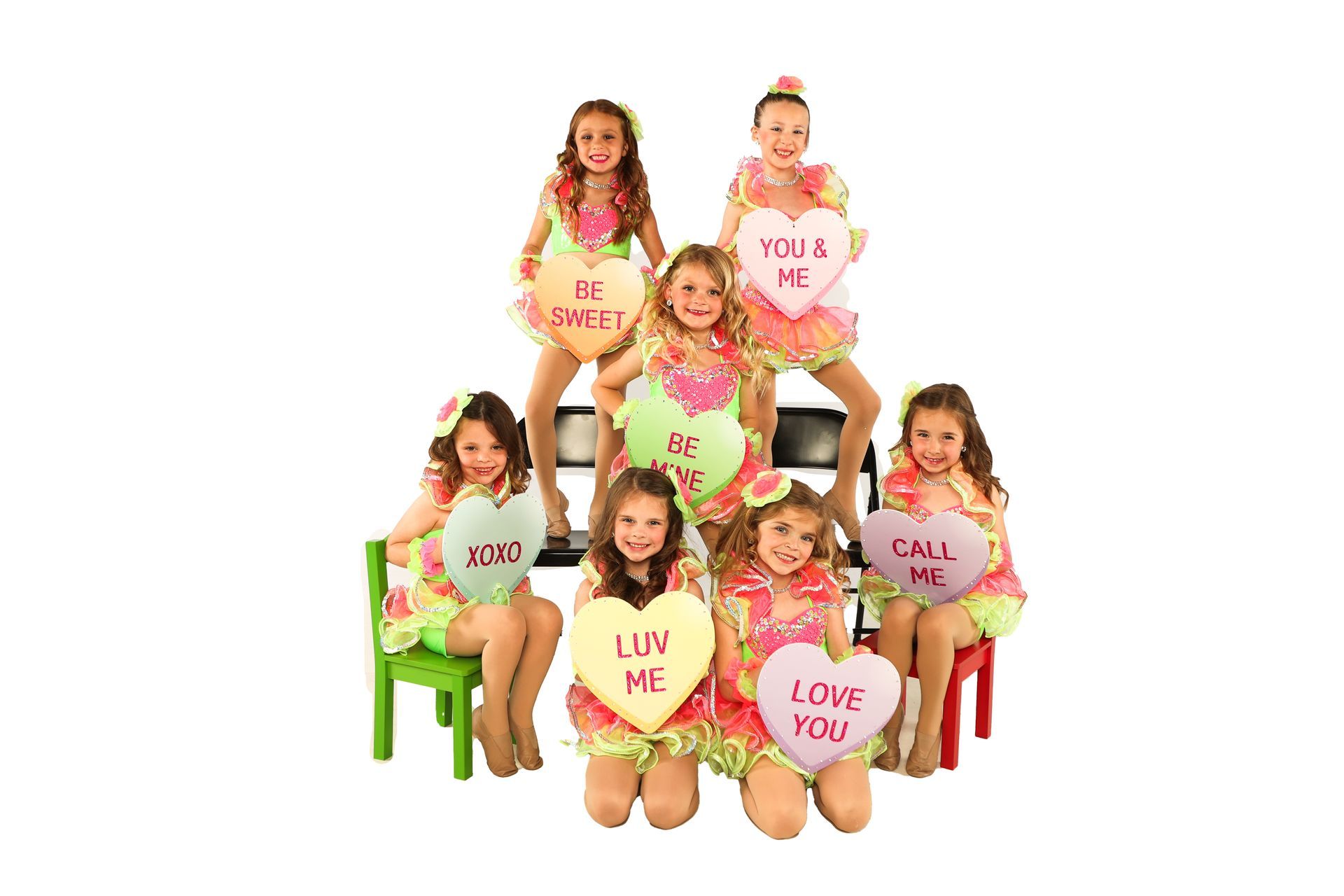 Five children seated on colorful chairs holding heart signs with loving messages against a white background