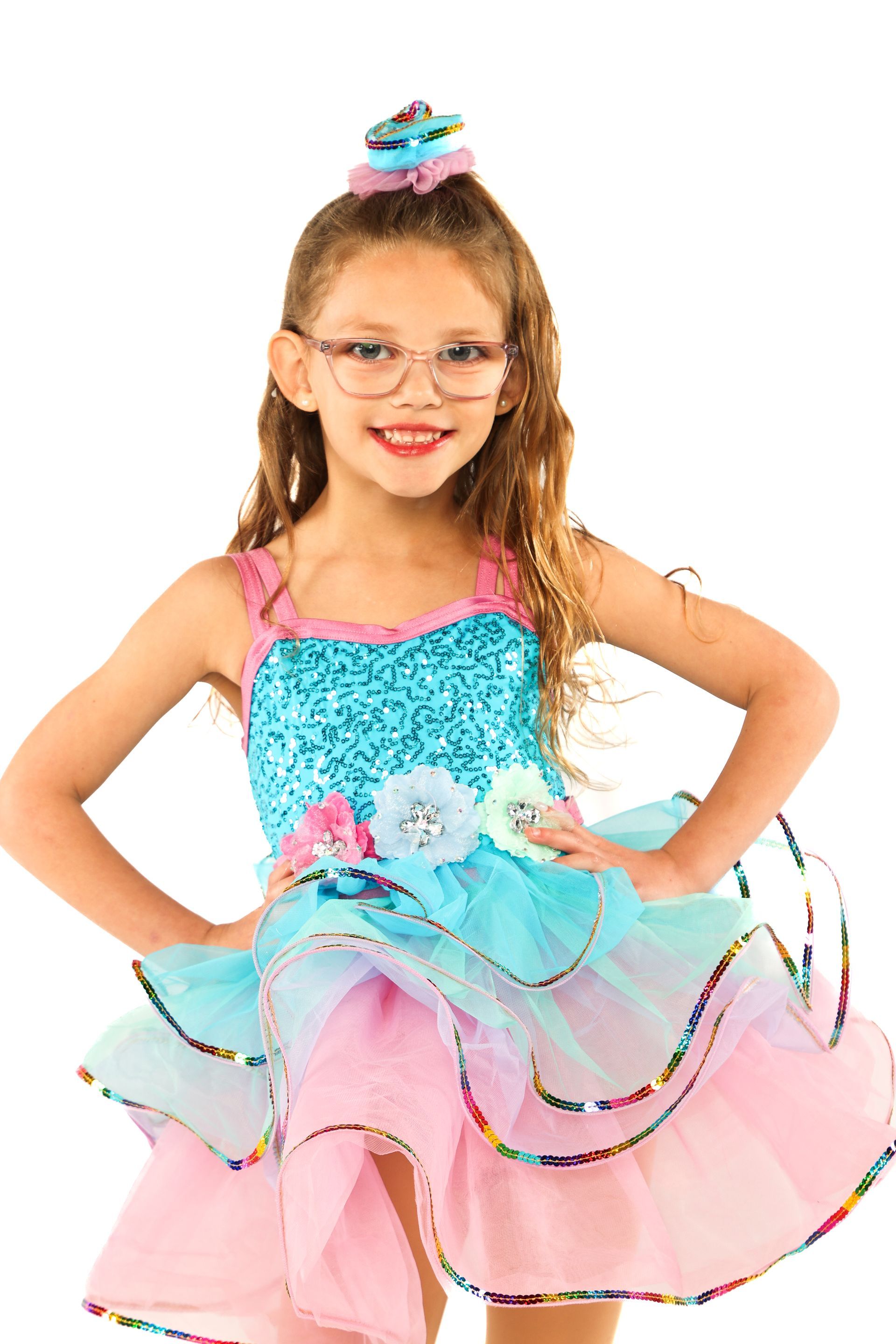 Young girl in a colorful tutu dress and tiara, smiling with hands on hips against a white background