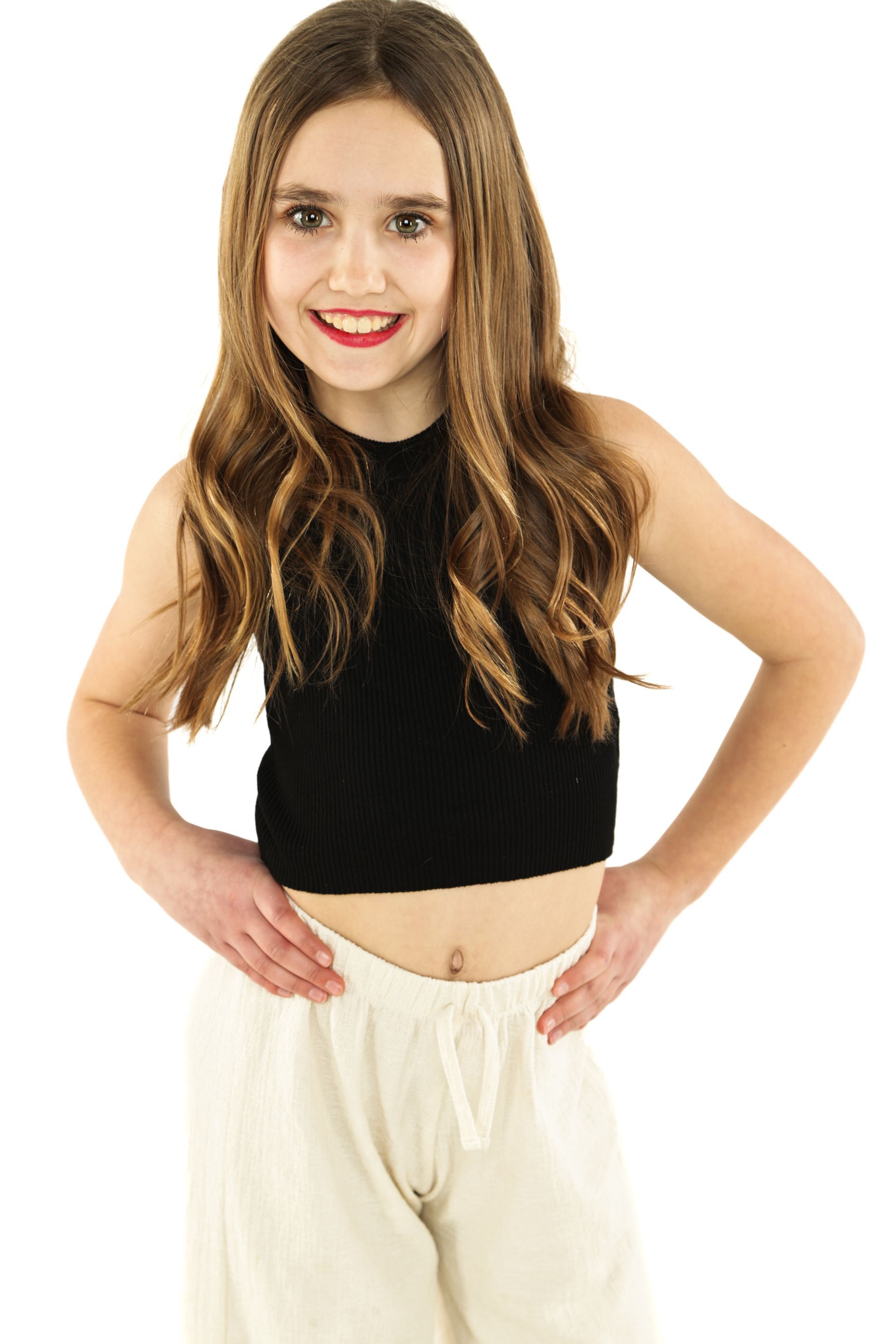Young woman standing against a white background, wearing a black crop top and cream pants, smiling.
