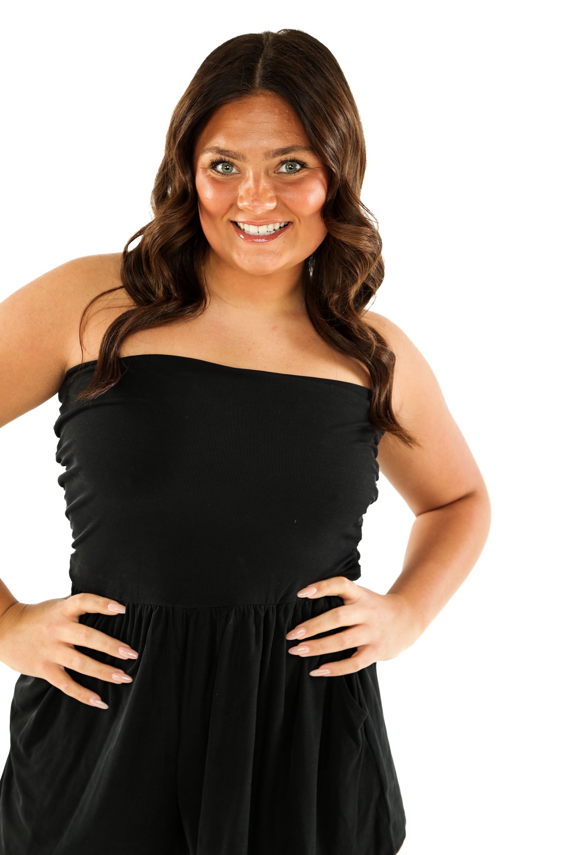 Woman in a black strapless dress smiling with hands on hips against a white background