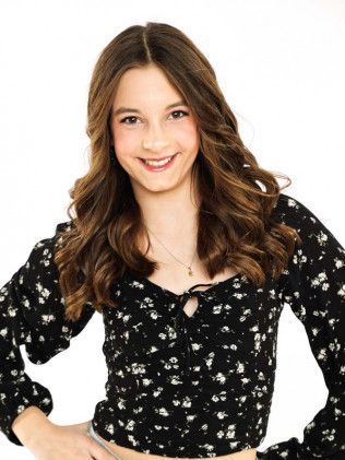 Smiling woman in a black floral blouse with wavy brown hair, standing against a white background