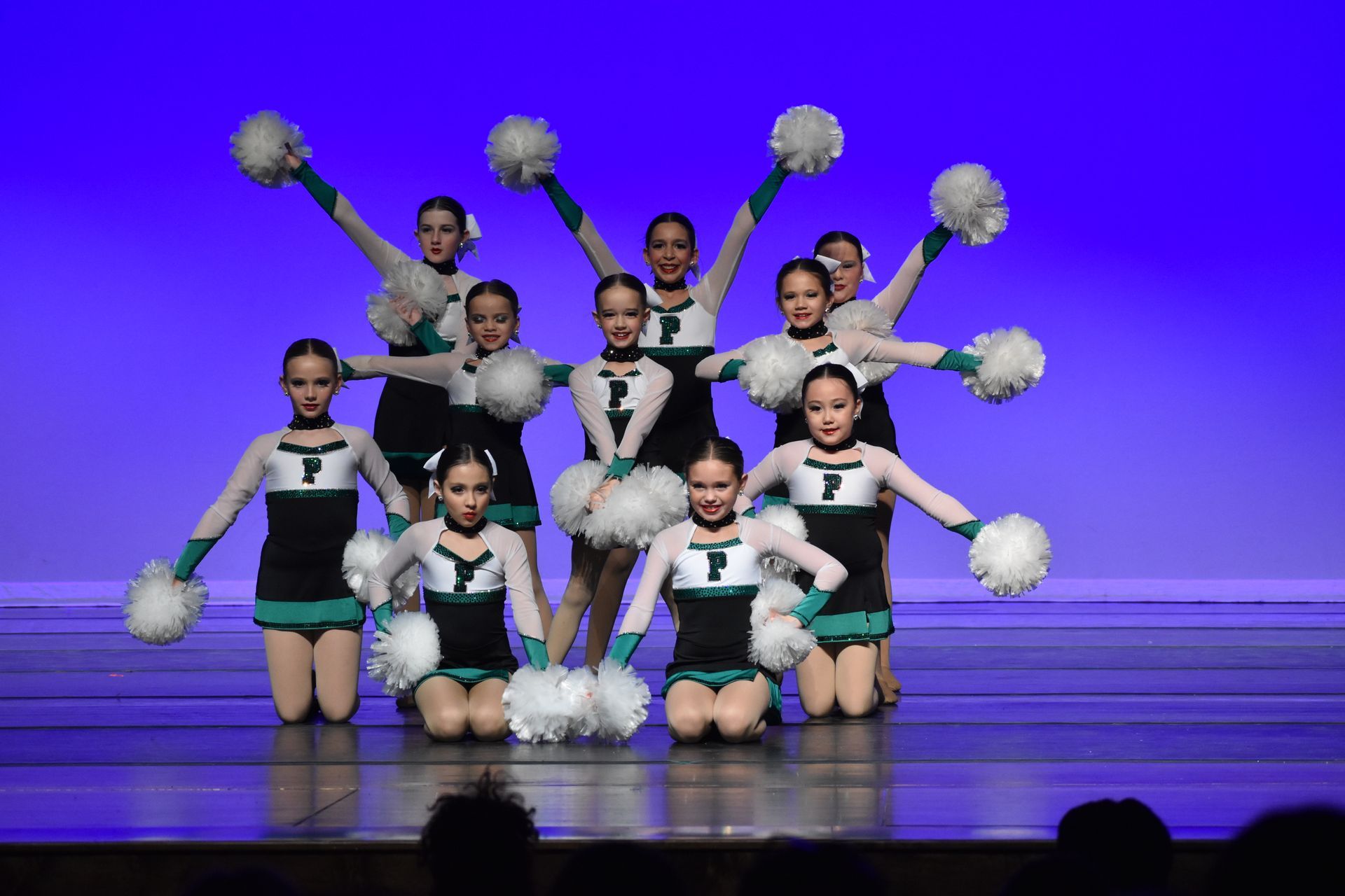 Cheer squad posing on stage with white pom-poms and teal-and-black uniforms under blue lighting