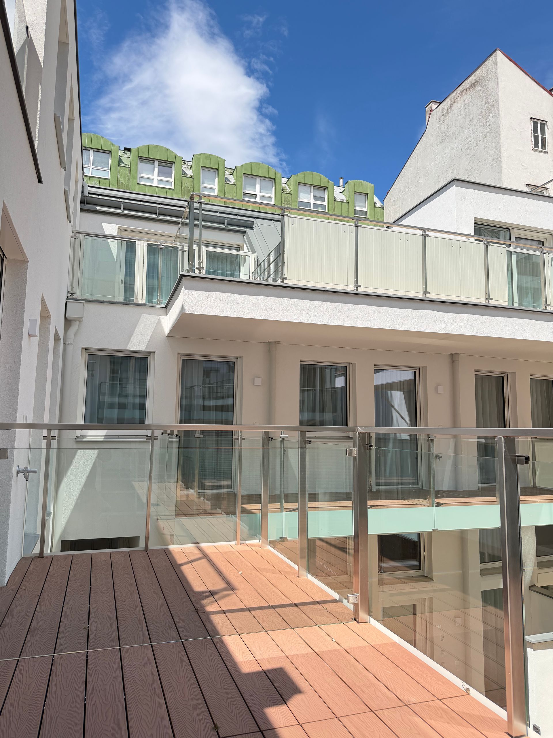 Sunny rooftop terrace with glass railing, wood deck, and white buildings under a blue sky