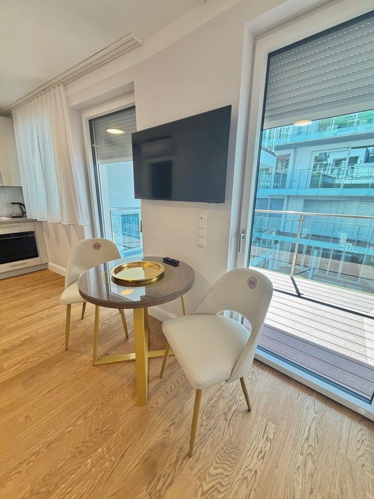 Modern living room with round table, two white chairs, wall-mounted TV, and large windows overlooking a balcony