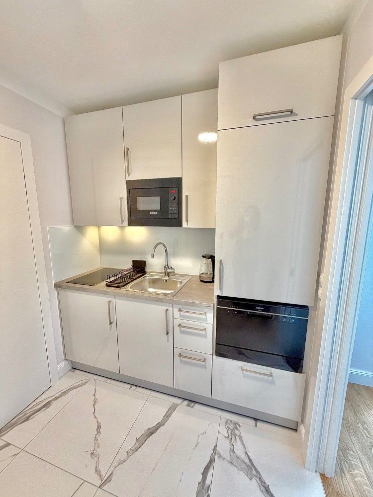 Small white kitchenette with built-in oven, stovetop, sink, and tall refrigerator in a bright room