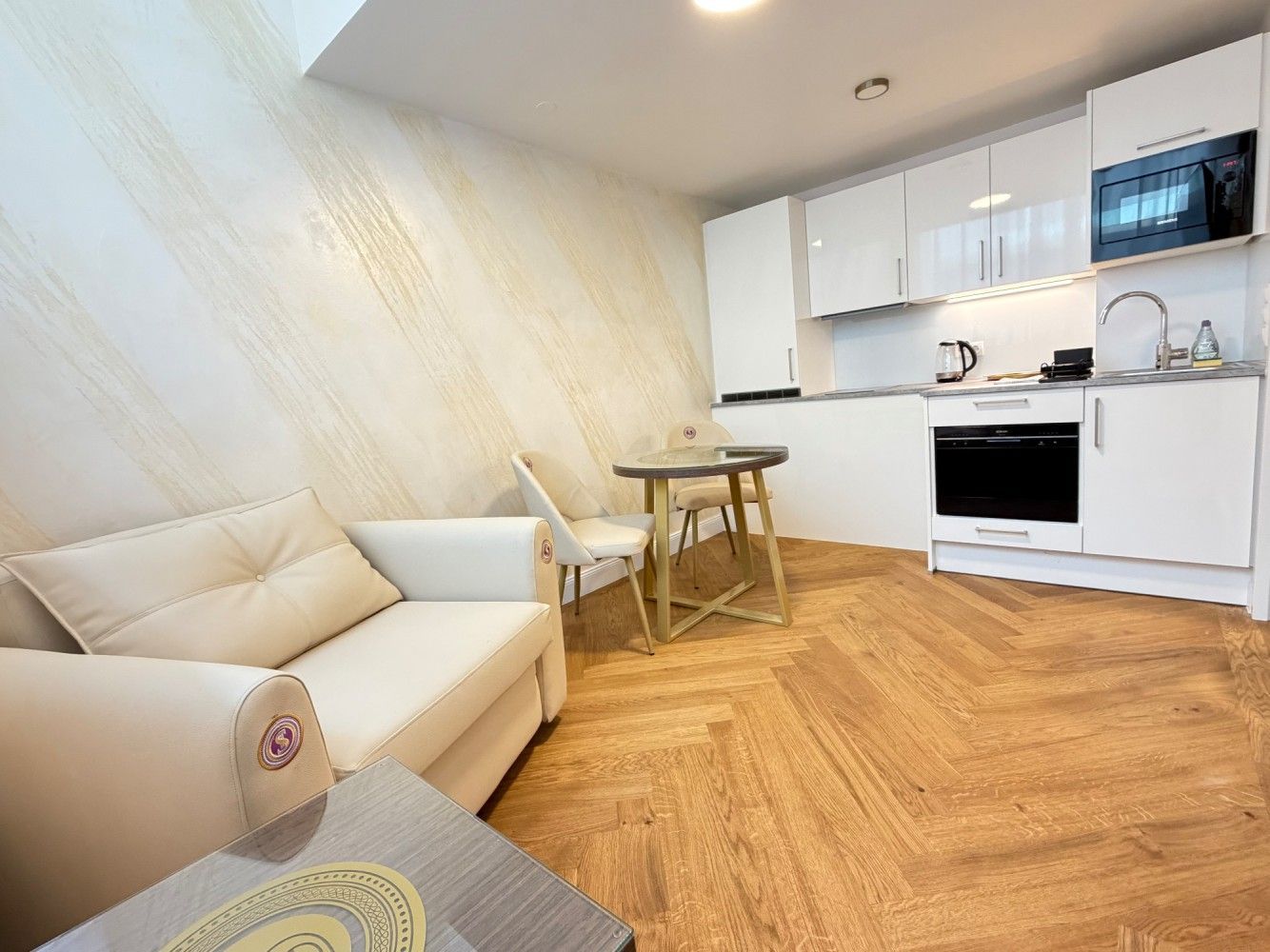 Bright modern studio apartment with beige sofa, small table, white kitchenette, and wood herringbone floor.