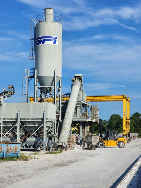 Industrial cement plant with tall silos, conveyor equipment, and a yellow loader beside a gravel road.